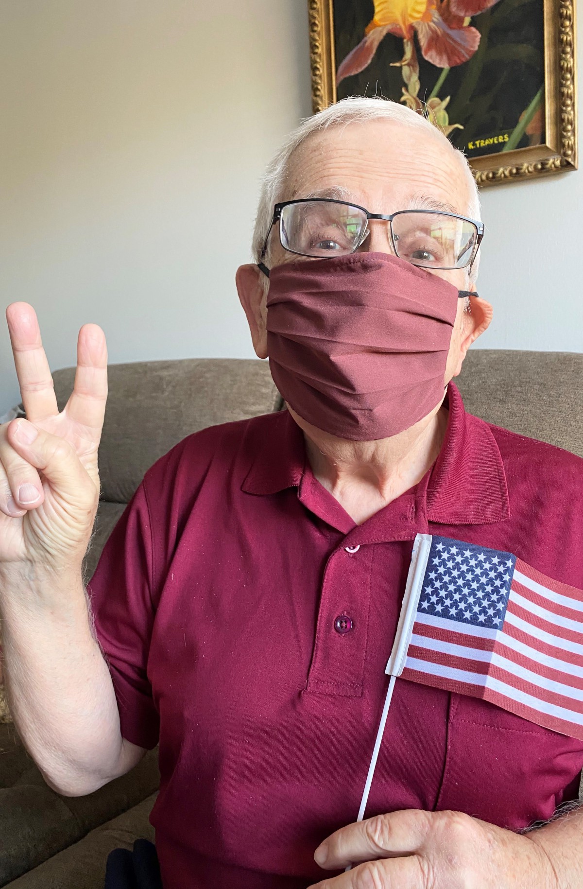 An elderly person wearing a face mask flashes a peace sign while holding an American flag indoors.