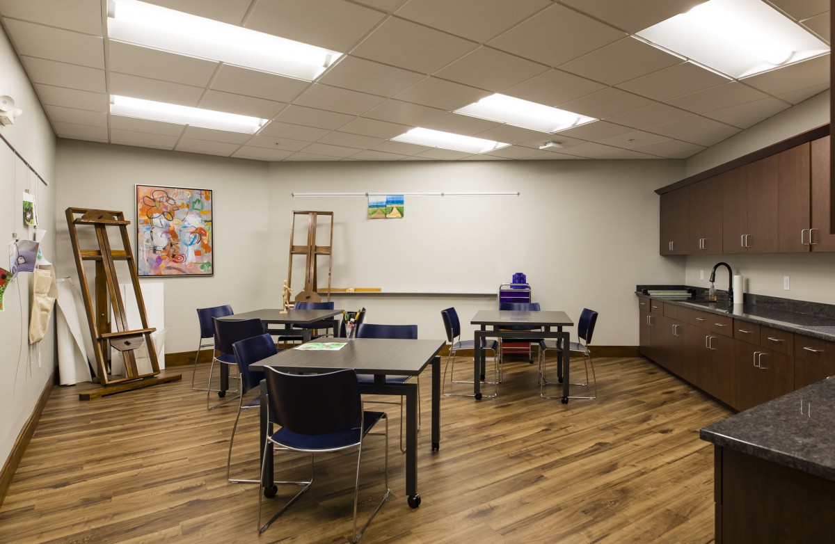 A well-lit classroom with tables, chairs, a whiteboard, an easel, and art supplies.