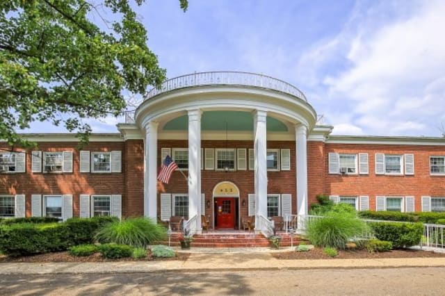 A grand mansion with large white columns and a red door, set amidst lush greenery.