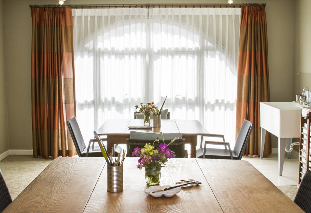 A sunlit dining area with a wooden table, chairs, and vases of flowers by the window.