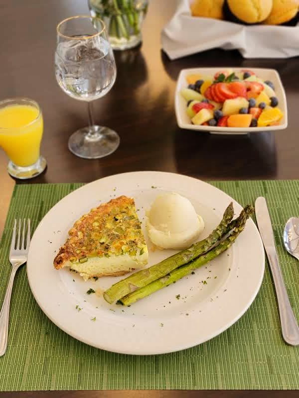 A breakfast meal featuring quiche, asparagus, and mashed potatoes with a side of fruit salad, served with orange juice and water.