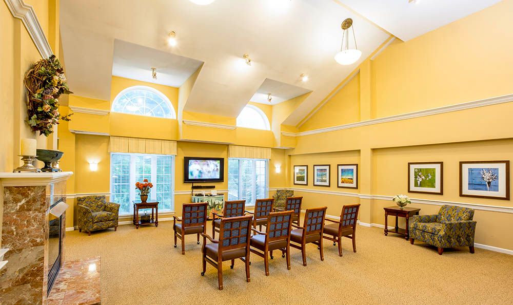 A brightly lit living room with a row of chairs facing a television, framed by large windows and yellow walls.