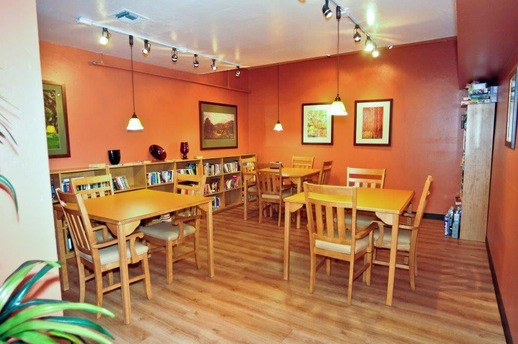 A cozy cafe interior with wooden tables and chairs, decorated with artwork and a bookshelf.