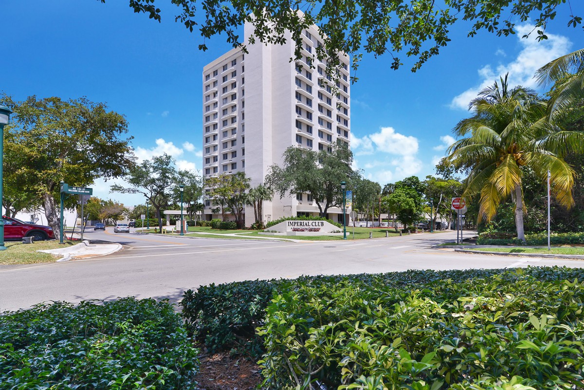 A tall, white high-rise building surrounded by lush greenery and trees under a bright blue sky.