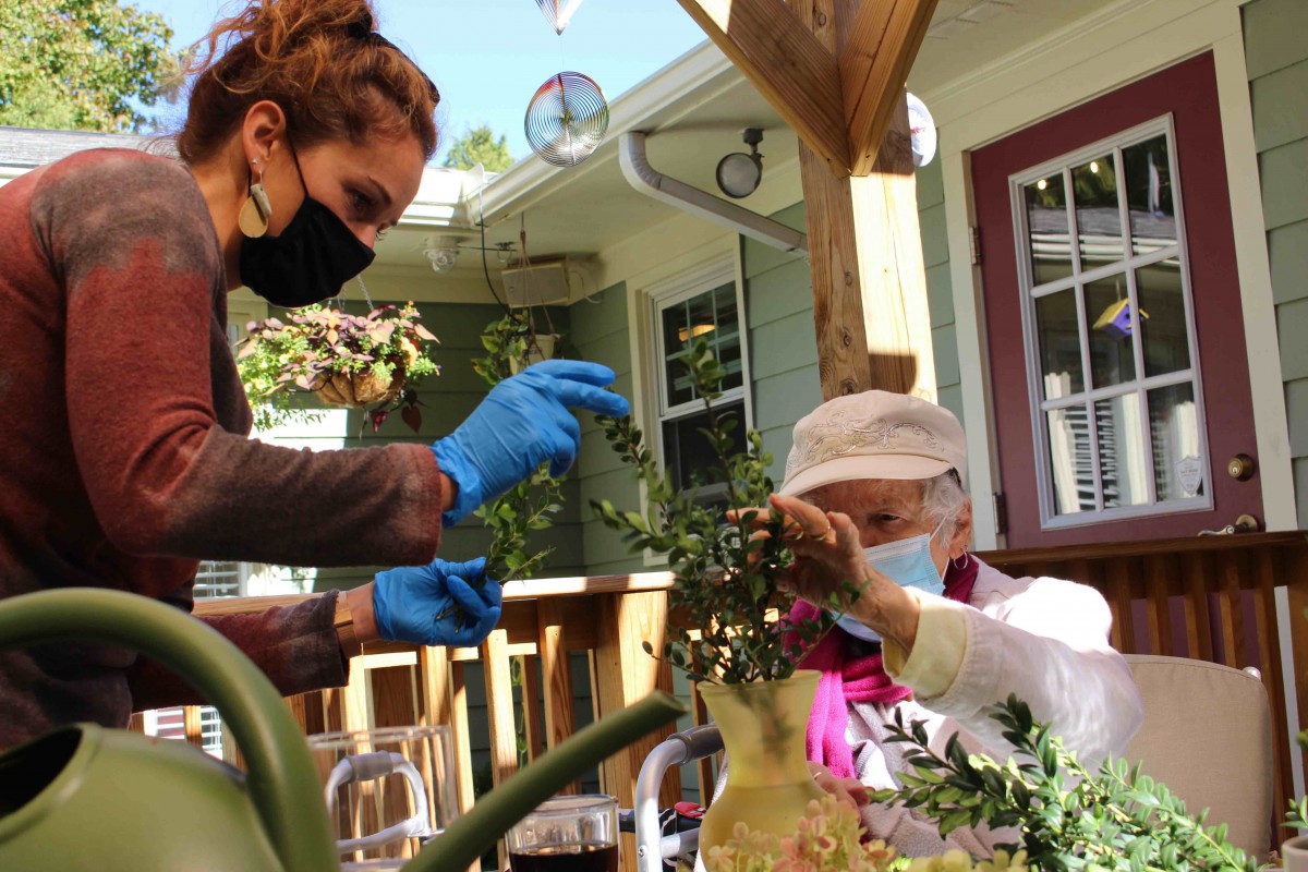 A caregiver assists an elderly person with gardening outdoors, both wearing face masks and gloves.