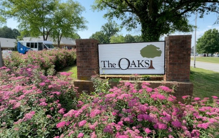A scenic view of a sign for 'The Oaks' surrounded by vibrant flowers and trees.