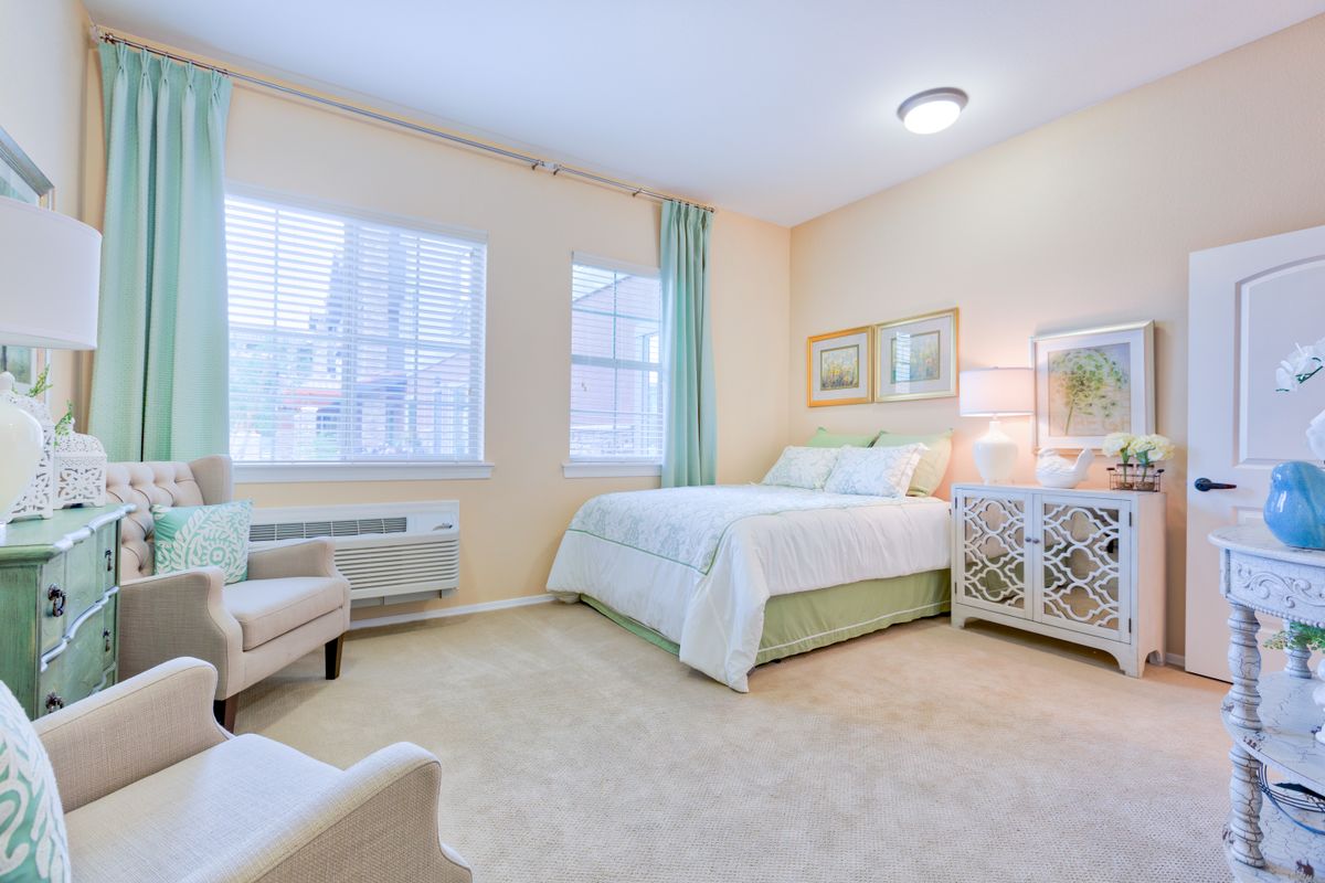 A cozy and elegantly decorated bedroom with light-colored walls, featuring a comfortable bed, chair, and windows with natural light.