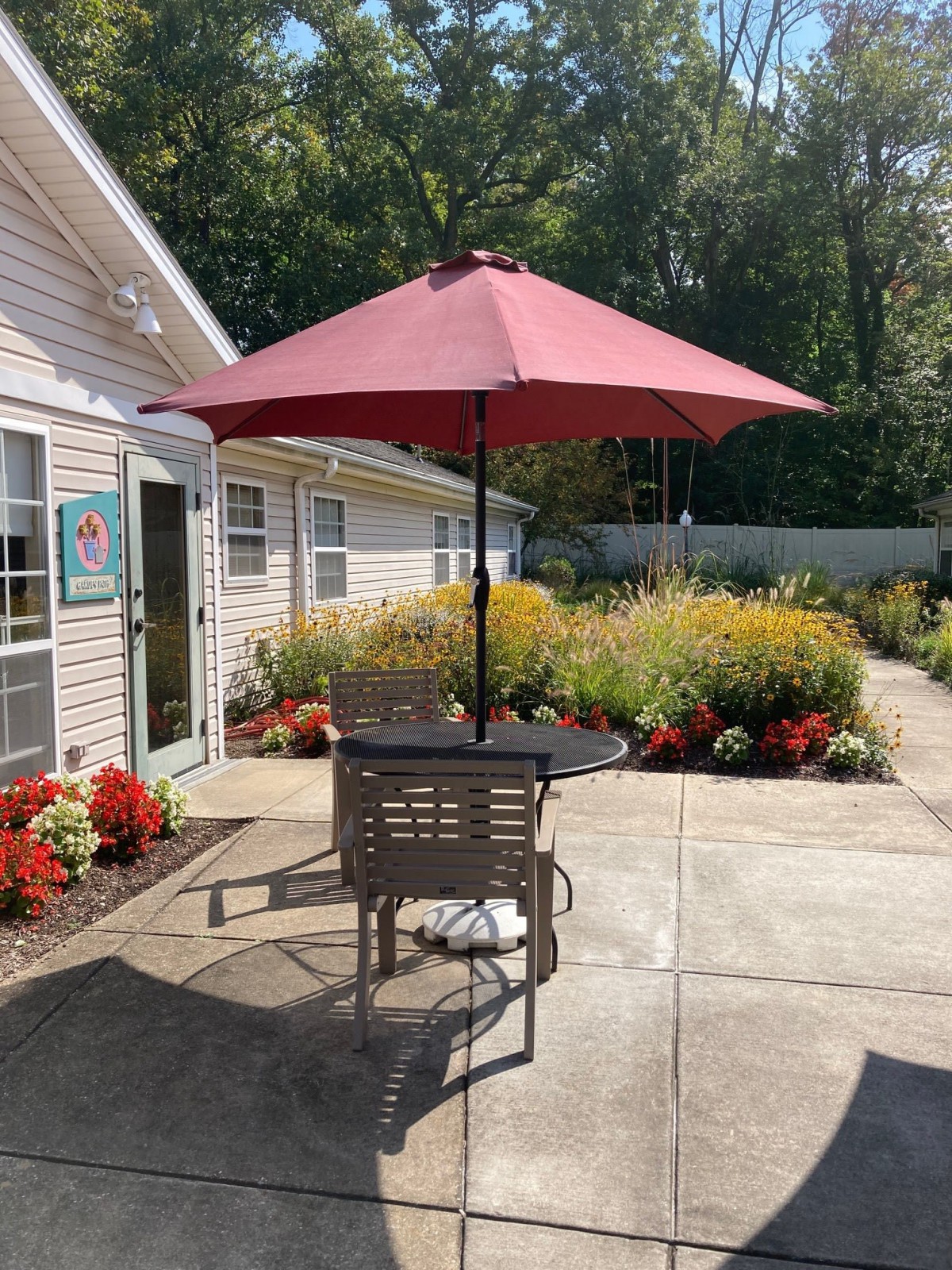 A sunny patio with a table and chairs under a red umbrella, surrounded by vibrant flowers.