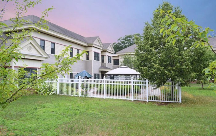 A view of a two-story residential building with a well-maintained lawn and white fence, featuring outdoor furniture under umbrellas.
