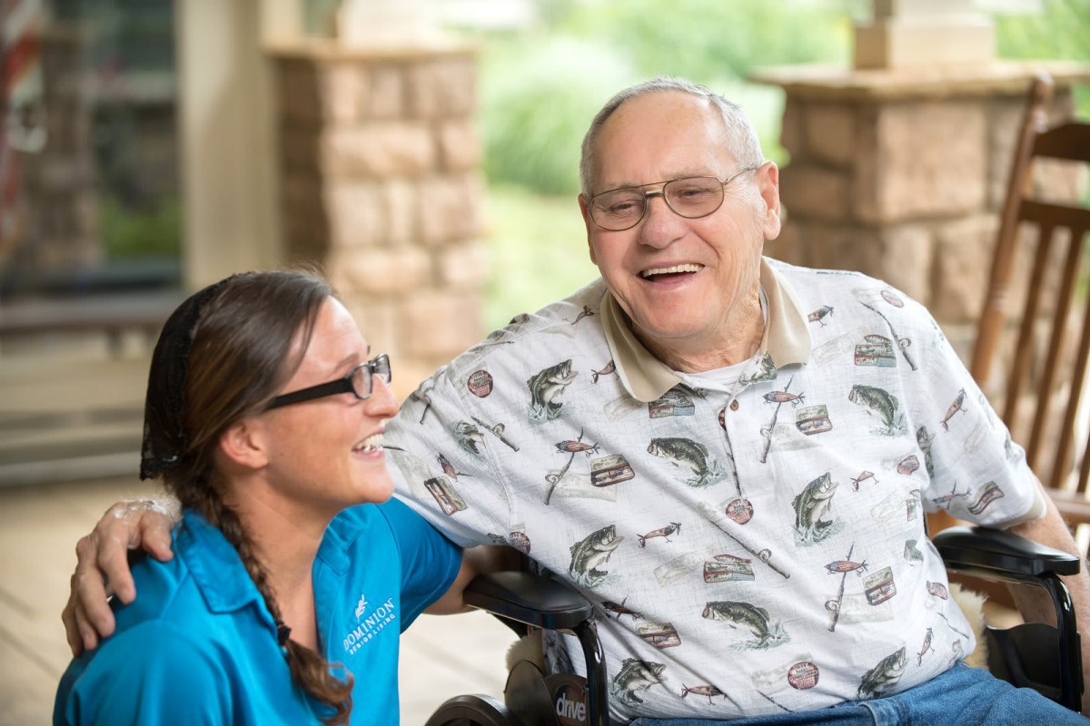 An elderly man in a wheelchair sharing a joyful moment with his caregiver outdoors.