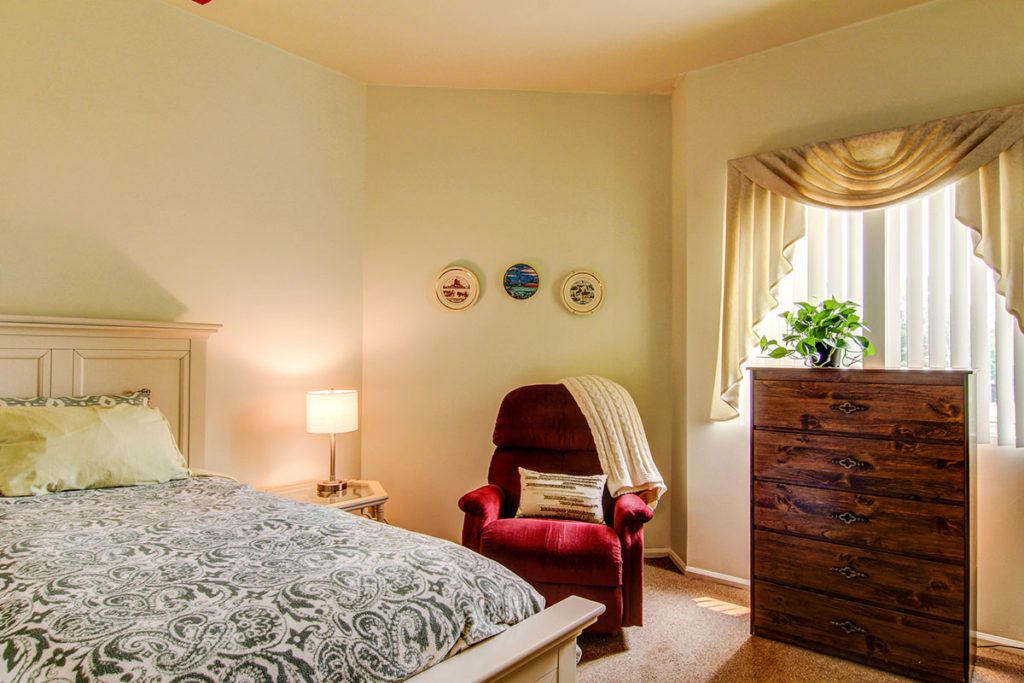 A cozy bedroom with a patterned bedspread, red armchair, wooden dresser, and soft lighting.