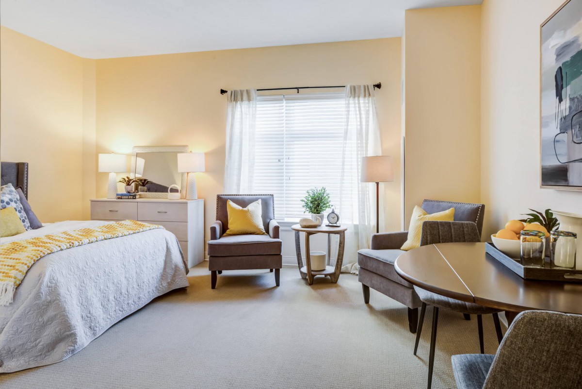 A cozy bedroom featuring a comfortable bed, two armchairs, and a small table near a window with sheer curtains.