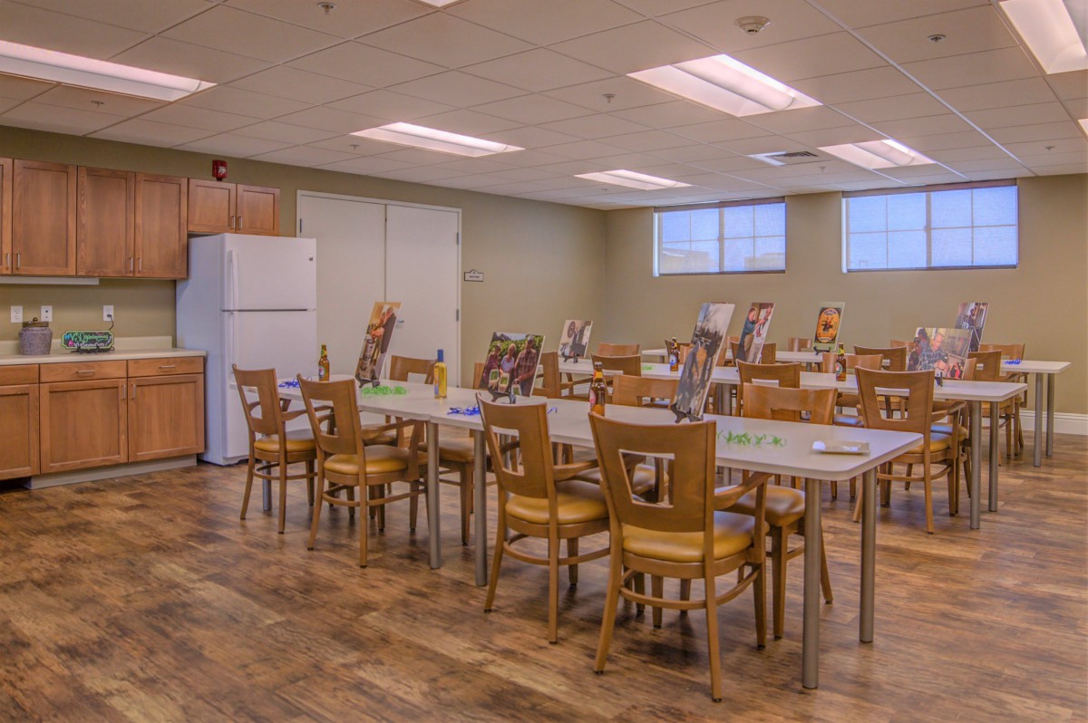 A well-lit dining room with several wooden tables and chairs, featuring a refrigerator in the corner.
