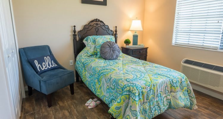 A cozy bedroom featuring a single bed with colorful bedding, a nightstand with a lamp, and an upholstered chair.