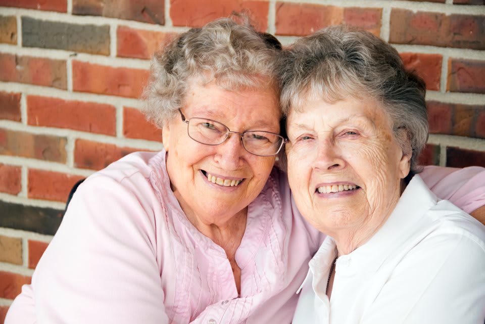 Two elderly women smiling and hugging in front of a brick wall.