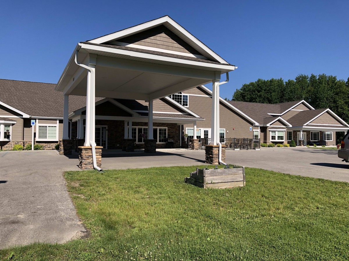 A building entrance with a canopy, surrounded by a driveway and grassy lawn on a sunny day.