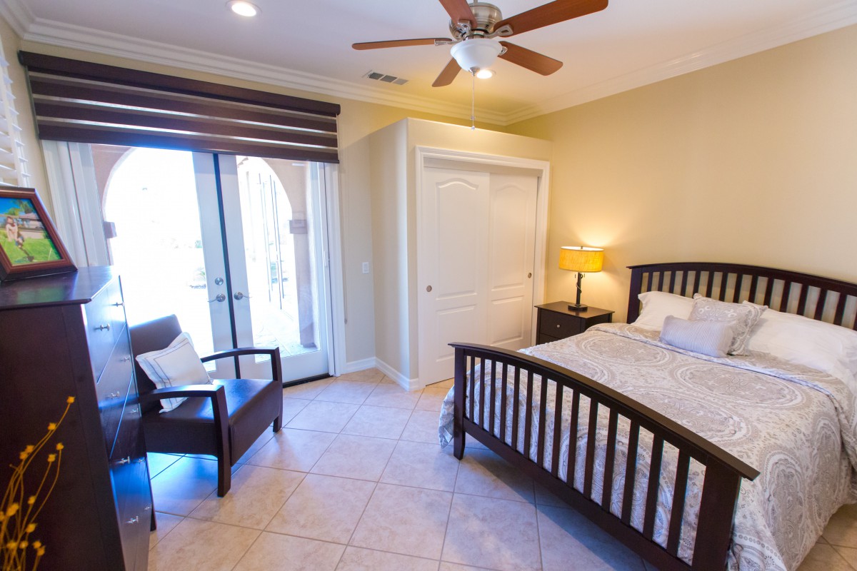 A cozy bedroom with a double bed, chair, lamp, and ceiling fan, featuring natural light from large windows.