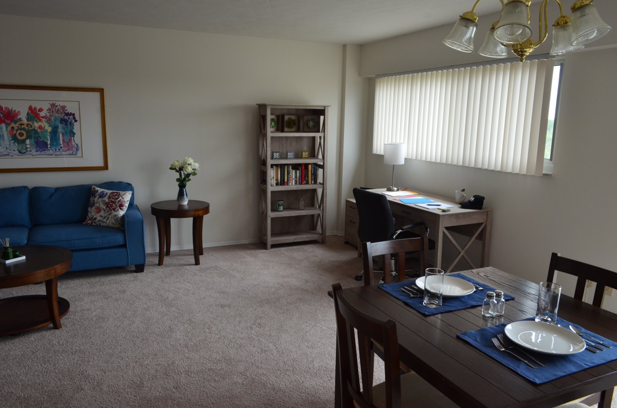 A cozy living and dining space featuring a bookshelf, desk with a lamp, dining table set for two, and comfortable seating.