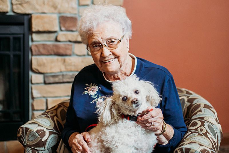 An elderly woman sitting indoors, smiling and holding a small dog.