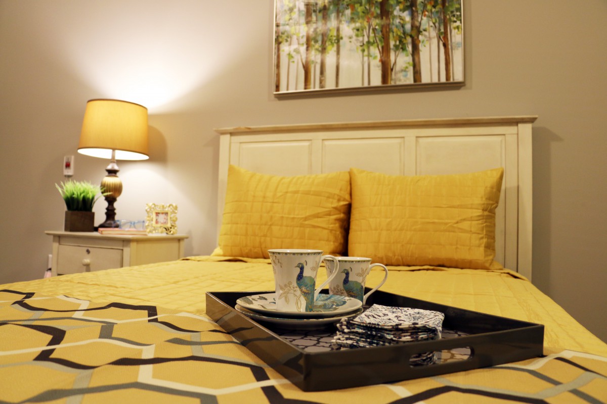 A cozy bedroom with yellow bedding and decorative pillows, featuring a tray with teacups on the bed and a lamp softly illuminating the room.