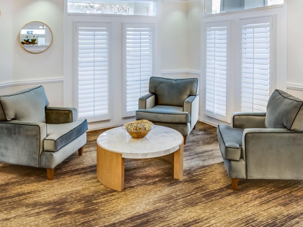 A cozy living room with three armchairs arranged around a round wooden coffee table, featuring large windows with shutters and a warm hardwood floor.