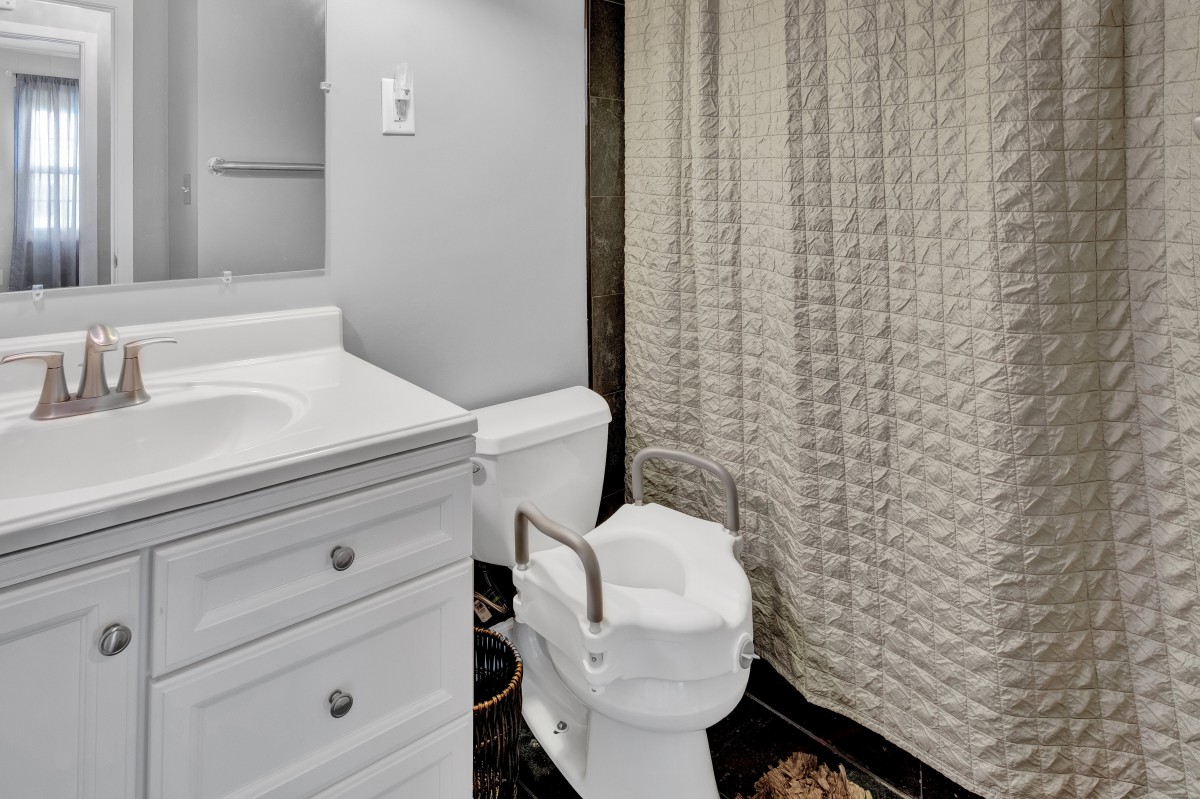 A bathroom featuring a toilet with a handrail for accessibility, a sink, and a patterned shower curtain.