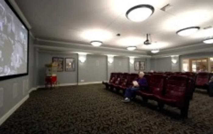 A small theater room with red seating and a single person sitting, illuminated by ceiling lights.