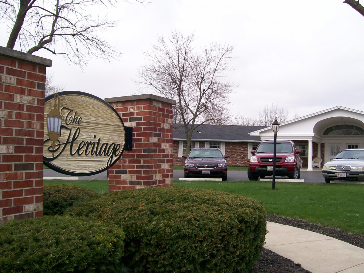 A view of a brick sign labeled 'The Heritage' in front of a building with two cars parked nearby.