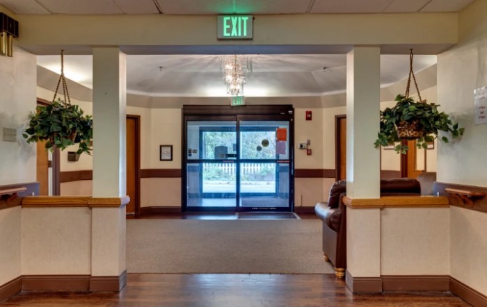 A well-lit hallway with an exit sign and double doors at the end, featuring decorative plants and seating.