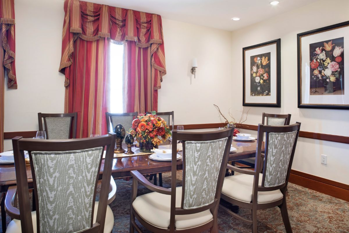 A cozy dining room with a wooden table, surrounded by upholstered chairs, adorned with red curtains and decorative items.