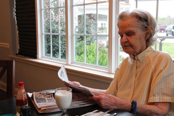 An elderly person sitting at a table by the window, reading a newspaper with a glass of milk nearby.