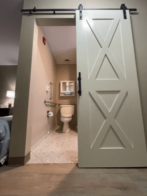 A modern bathroom with a sliding barn door and a tiled floor, adjacent to a hotel room.