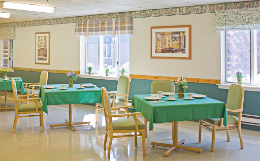 A bright and inviting dining area featuring tables with green tablecloths, comfortable chairs, and floral arrangements near the windows.