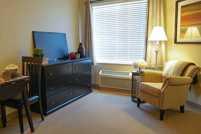 A cozy hotel room with a comfortable chair, dresser, and window with blinds.