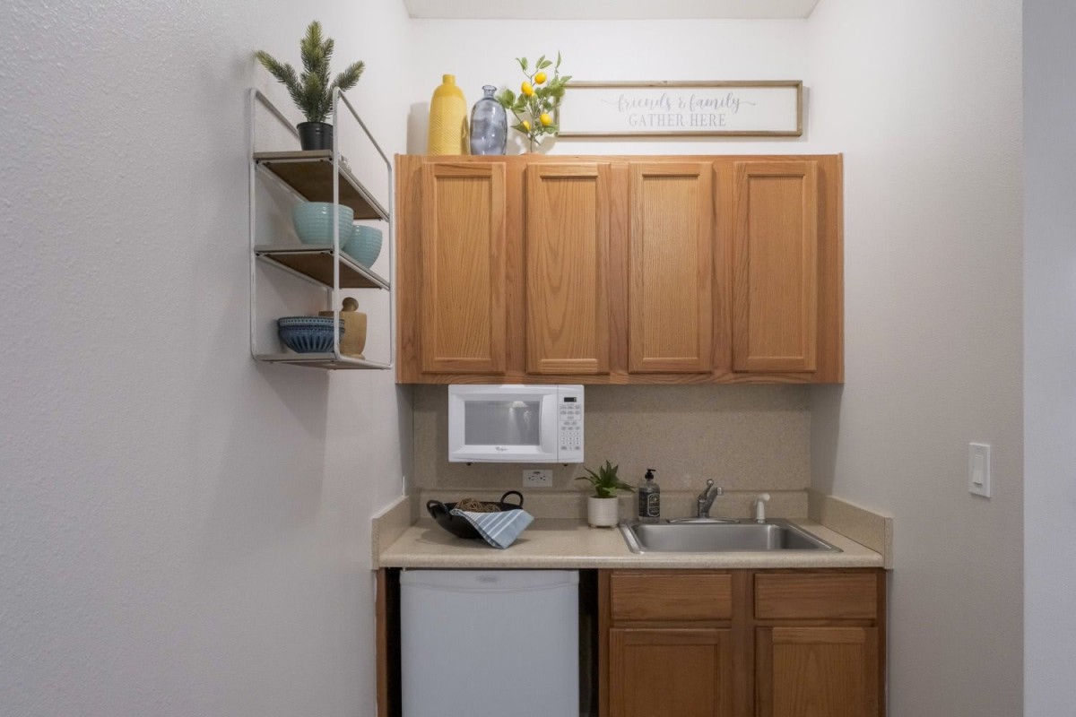 A compact kitchenette with wooden cabinets, a small sink, a microwave, and decorative shelves with plants.
