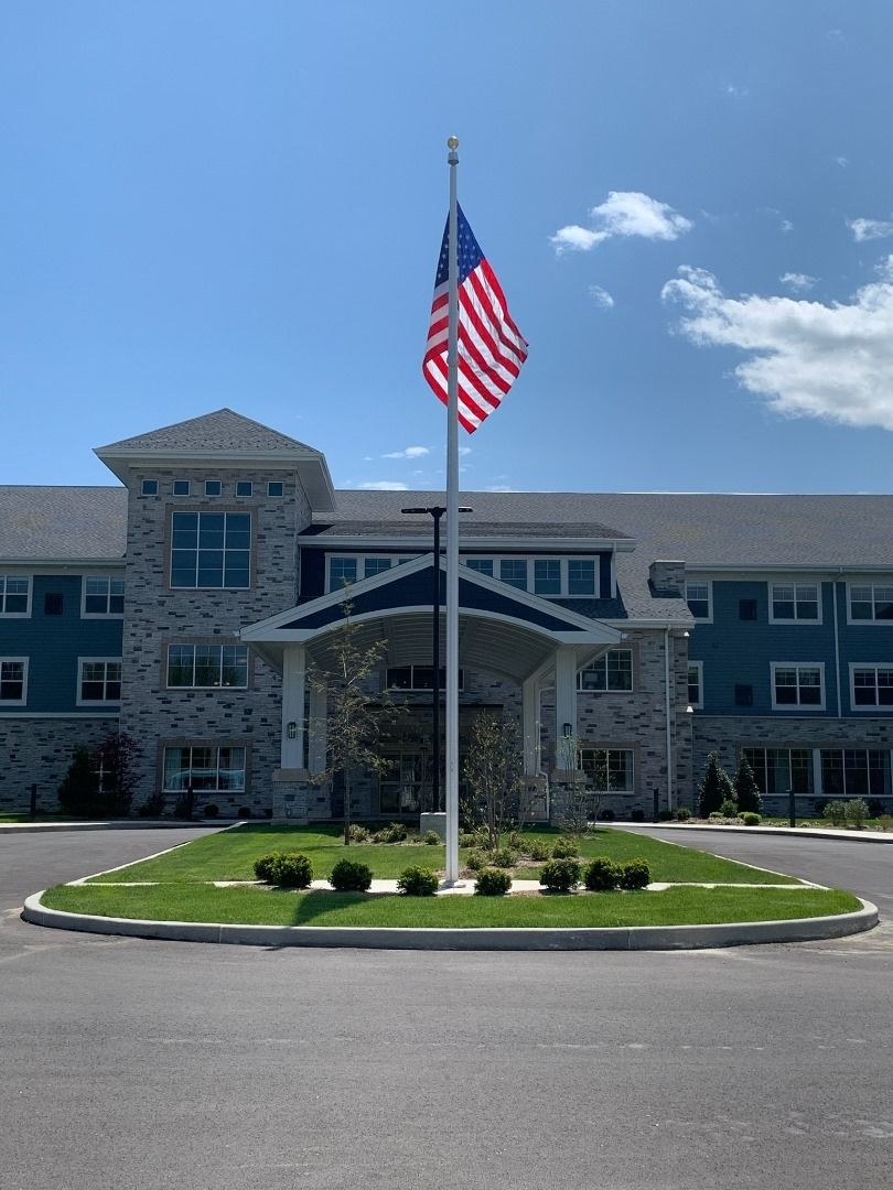 A large building with an American flag prominently displayed in front, surrounded by well-maintained landscaping and a clear blue sky.