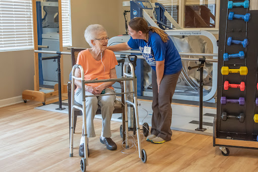 A caregiver assists an elderly woman using a walker in a rehabilitation setting.