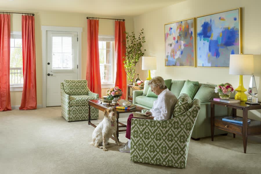 An elderly person sits in a cozy living room with their dog, surrounded by colorful decor and artwork.