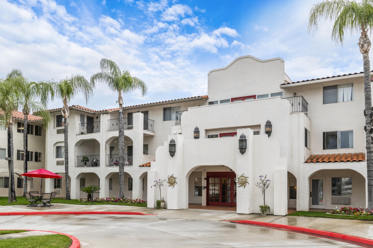 A Spanish-style residential building with palm trees and a partly cloudy sky.