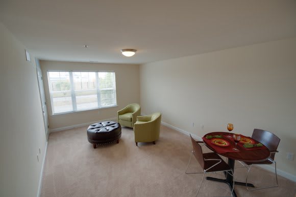 A cozy living room with two green chairs, a round ottoman, and a small dining table by the window.