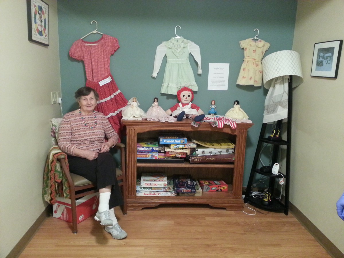 A woman seated beside a shelf displaying vintage dolls and board games with dresses hanging on the wall.