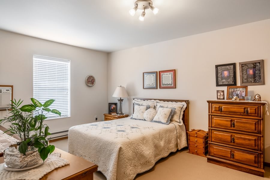 A cozy bedroom featuring a wooden bed with a light-colored quilt, matching dresser and nightstand, adorned with decorative wall art and a potted plant by the window.