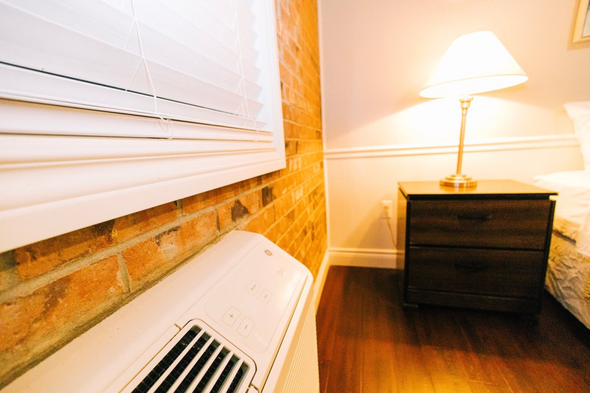 A cozy room setup featuring an air conditioner, a brick accent wall, and a warmly lit lamp on a nightstand.