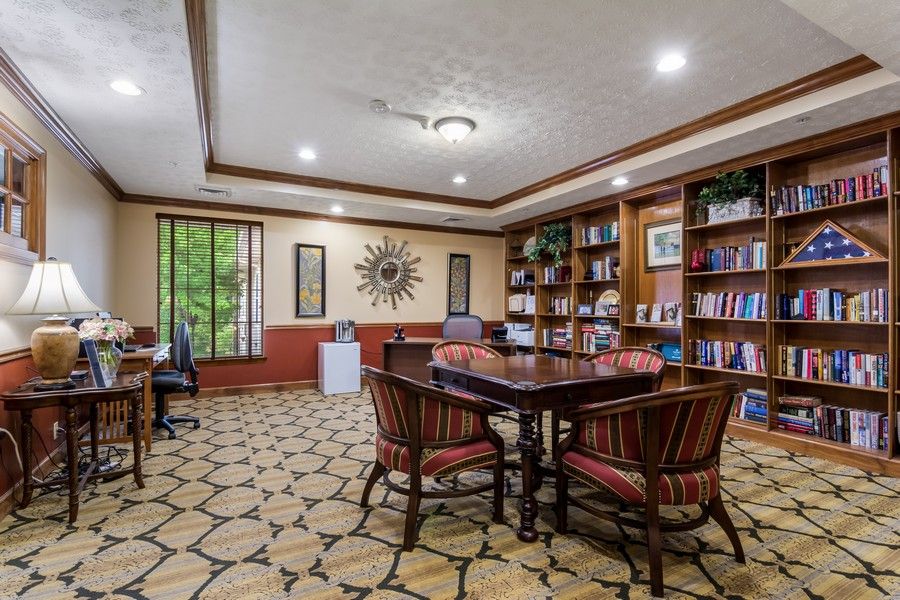 A cozy library room with bookshelves, a central table with chairs, and large windows providing natural light.