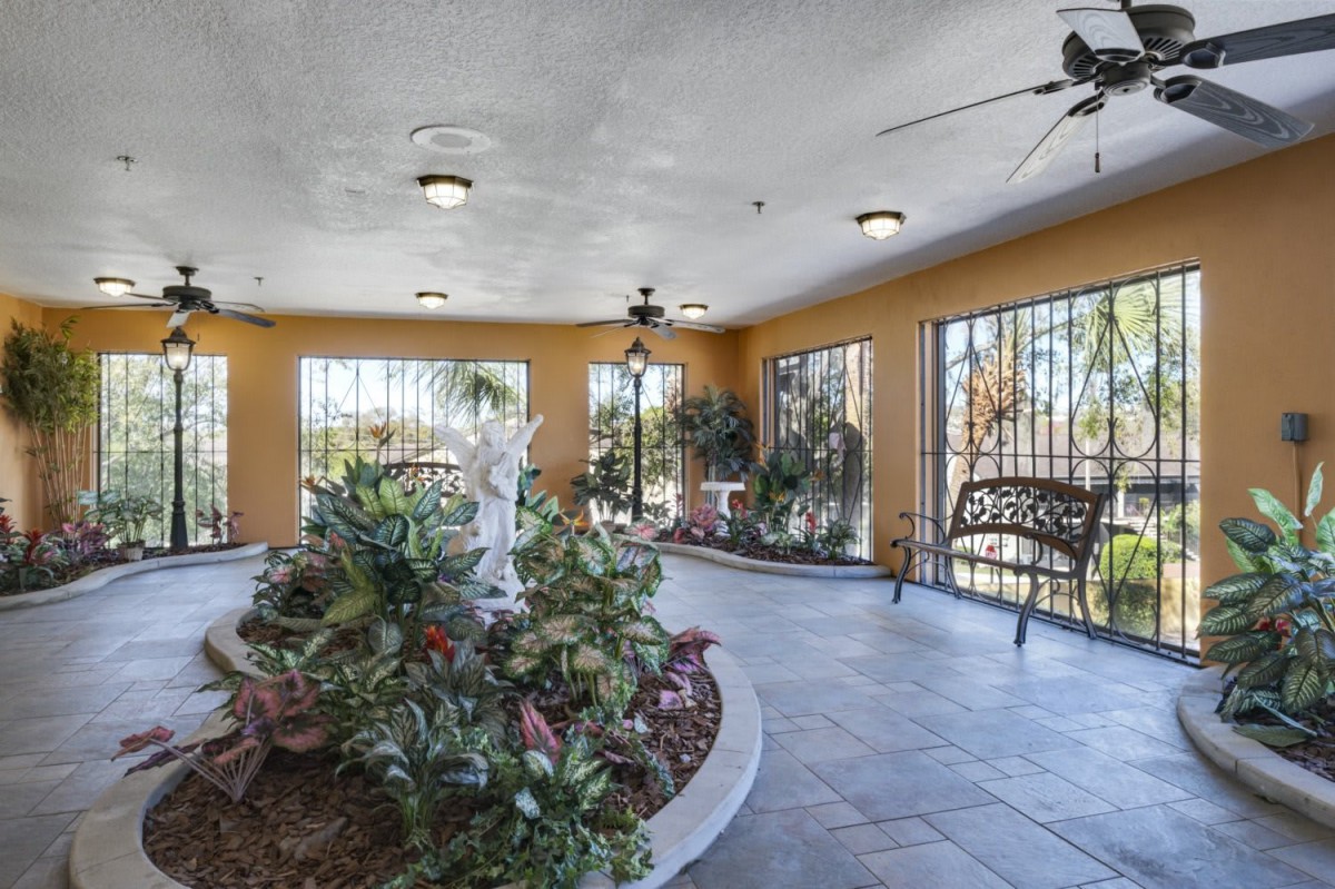 A bright indoor space featuring a garden with a statue, surrounded by large windows and ceiling fans.