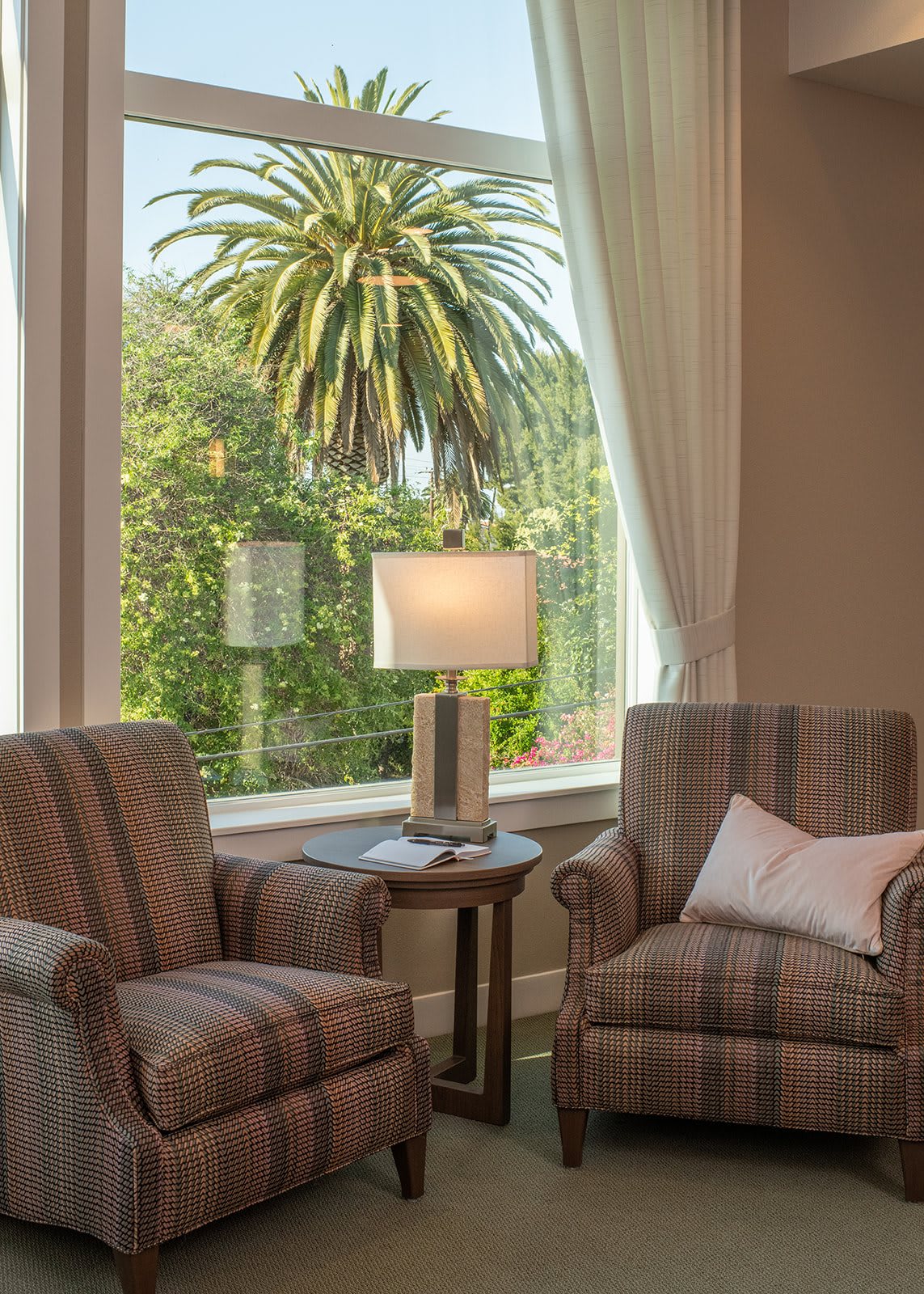 A cozy living room with two chairs and a lamp by a large window overlooking a palm tree.
