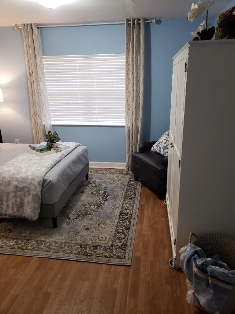 A cozy bedroom with a neatly arranged bed, wardrobe, armchair, and window with blinds.