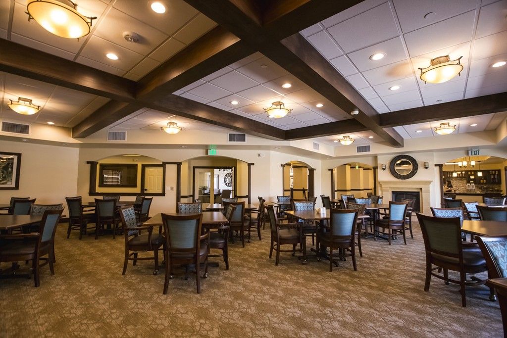 A spacious dining room with multiple tables and chairs, featuring warm lighting and a carpeted floor.