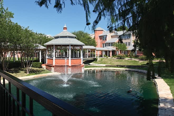 A serene outdoor scene featuring a gazebo by a pond with a fountain, set against the backdrop of a charming building.