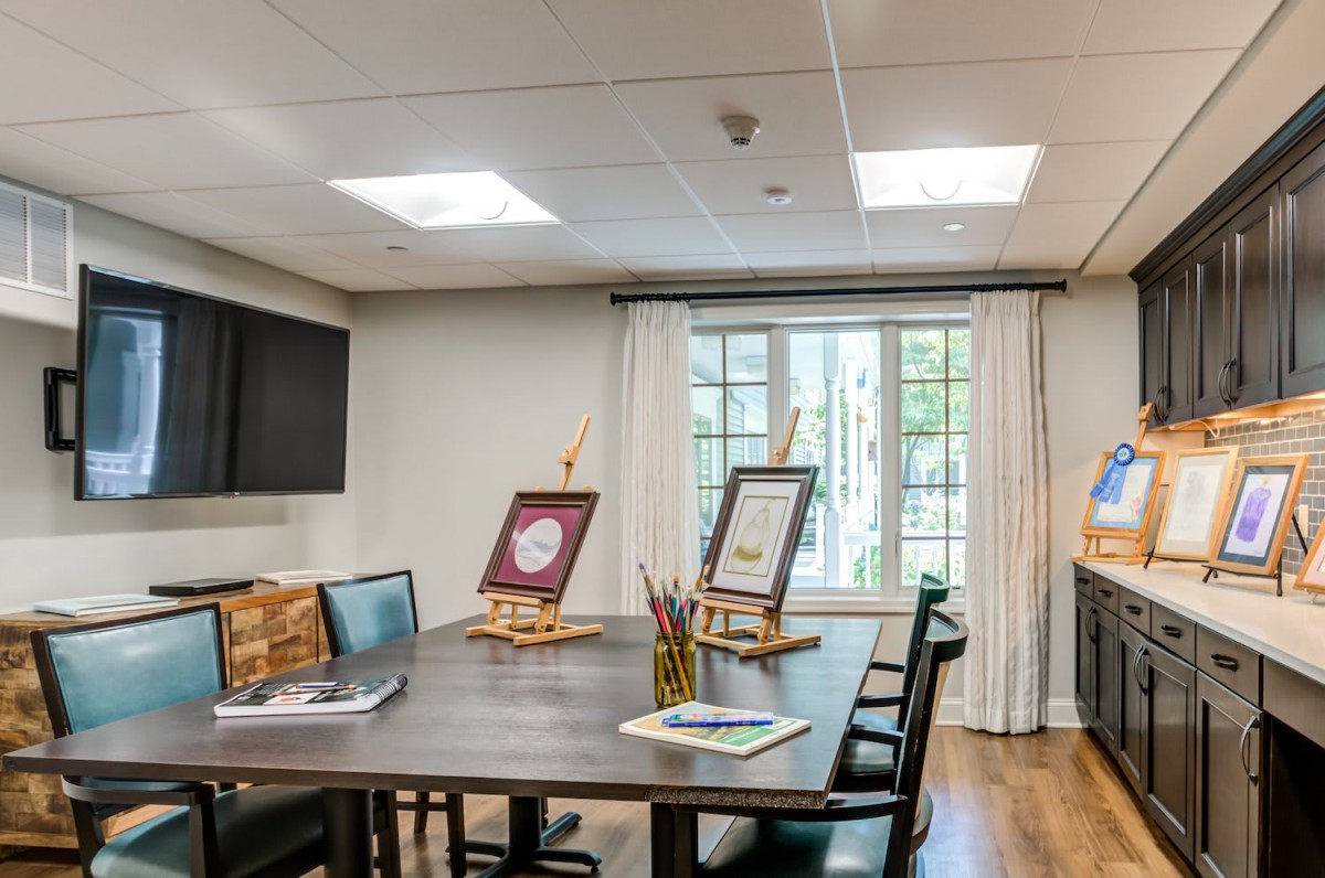 A bright art room with easels set up on a table, surrounded by chairs and illuminated by natural light from a large window.